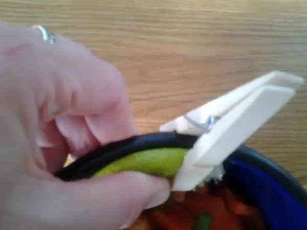 Hand hält Limettenscheibe mit Wäscheklammer über Schüssel mit geschnittenen Paprika auf Holztisch.