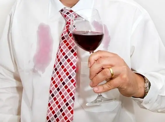 Ein Mann hält ein Glas Rotwein, während seine weiße Hemdbrust mit Flecken in einem hellen Raum markiert ist.