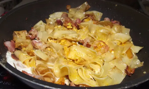 Gebratene Nudeln liegen in einer Pfanne, während sie mit Speck und Gewürzen aromatisiert werden, um Reste aufzuwerten.