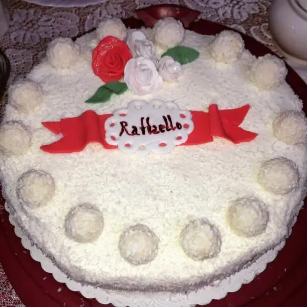 Eine Raffaello Torte mit weißen Kugeln und roten Zuckerdekoren thront auf einem Tisch, verziert mit "Raffaello" Schriftzug.