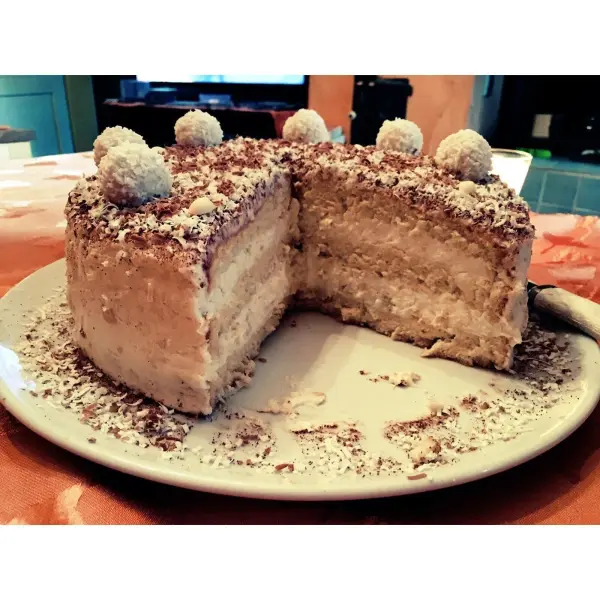 Eine Raffaello Torte auf einem Teller fehlt ein Stück, bestreut mit Kokos und Schokolade in einer gemütlichen Küchenszene.