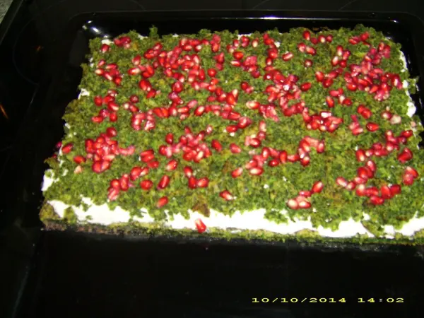 Ein grüner Mooskuchen mit scharlachroten Granatapfelkernen ist in einem dunklen Backblech angerichtet und dekoriert.