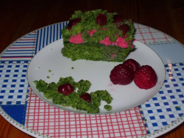 Ein grüner Mooskuchen liegt auf einem Teller, dekoriert mit Himbeeren, umgeben von buntem Geschirr.