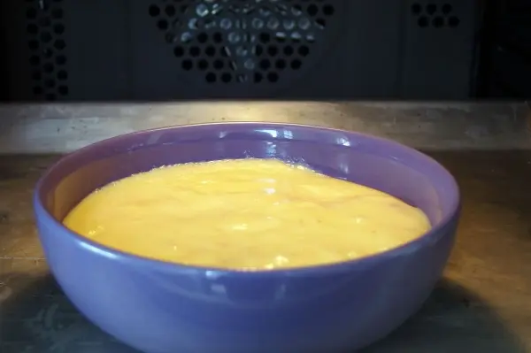 Eine lila Schüssel steht auf einem Tisch und enthält eine cremige Käsekuchenmasse, die im Ofen gebacken wird.