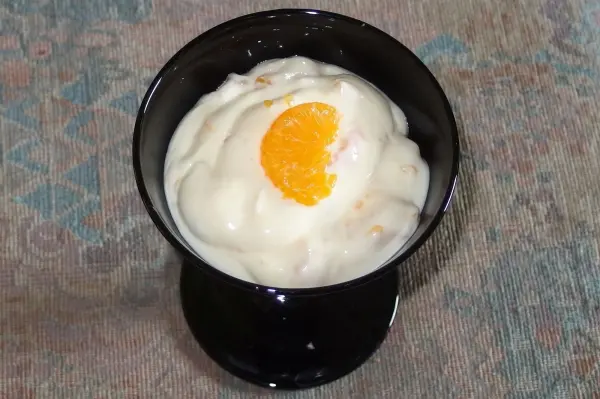 Ein schwarzes Dessertglas enthält cremigen, käsekuchenartigen Nachtisch, garniert mit einer halben Orange. Der Hintergrund zeigt eine strukturierte Oberfläche.