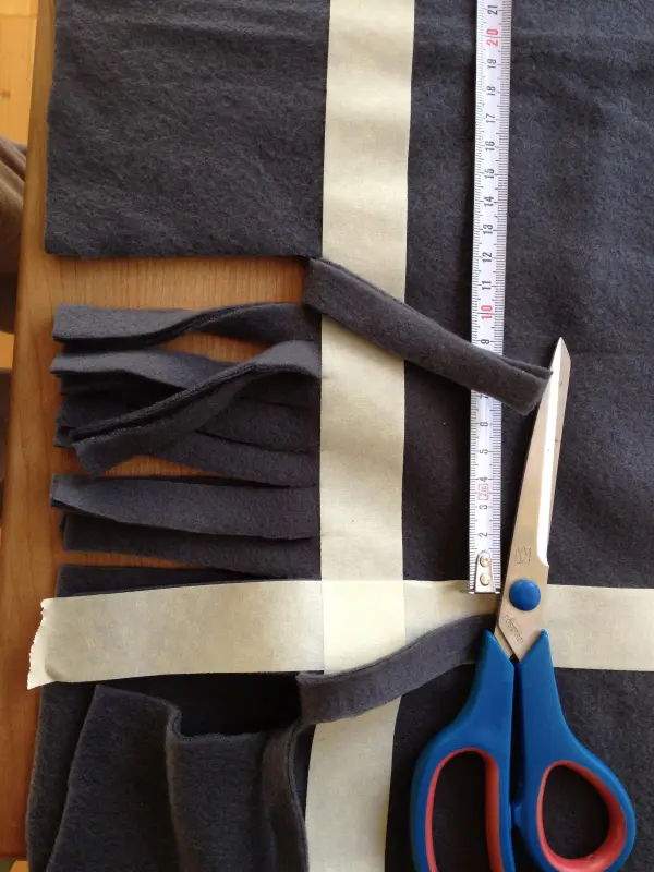 Eine Schere schneidet grauen Fleece-Stoff, während ein Maßband und Klebeband die Materialien für Kissen für Hund und Katze anordnen.