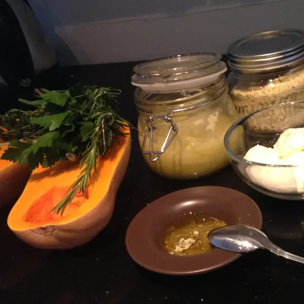 Ein halber gebackener Butternut-Kürbis liegt neben Kräutern und Zutaten für eine Kürbis-Kräutercreme auf einem dunklen Tisch.