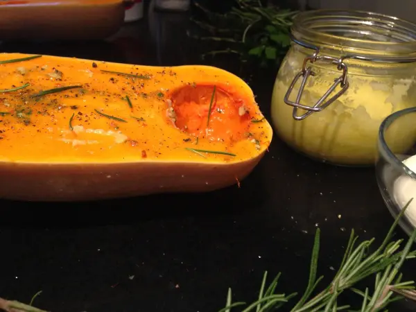 Gebackener Butternut-Kürbis liegt auf einem Tisch, umgeben von frischen Kräutern und einer Kräutercreme im Glas.