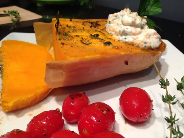 Der gebackene Butternut-Kürbis liegt auf einem Teller, garniert mit Kürbis-Kräutercreme und umgeben von roten Kirschtomaten.