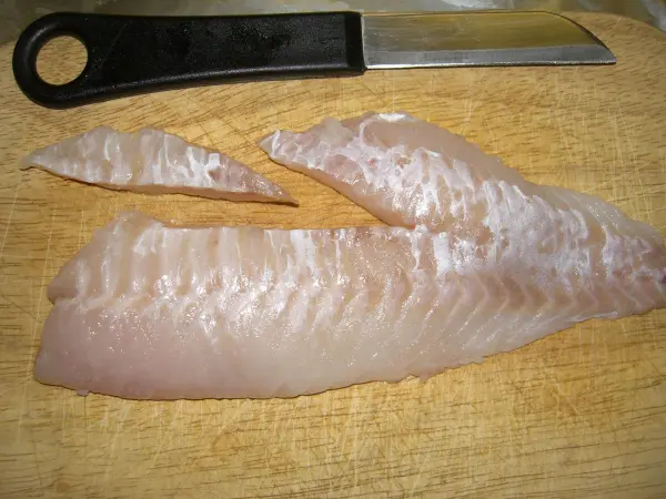 Ein Fischfilet liegt auf einem Holzbrett, neben einem Messer, bereit zum Entgräten in einer Küchenumgebung.