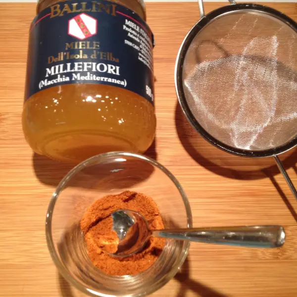 Ein Glas mit rotem Gewürz steht neben einem Honigglas und einem Sieb auf einem Holzbrett, bereit zum Vermischen.