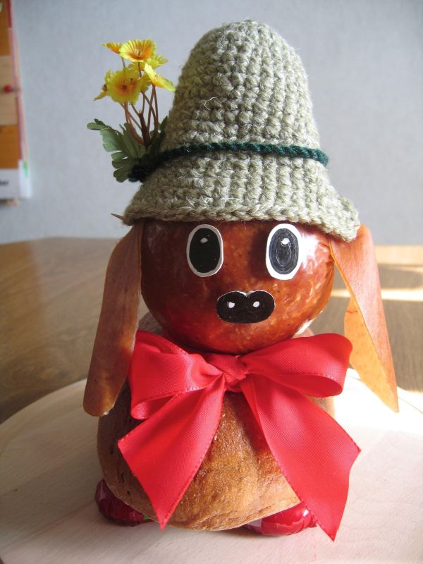 Ein Brotdackel steht auf einem Tisch, trägt einen gehäkelten Hut und eine Schleife und hat Blumen im Kopf.