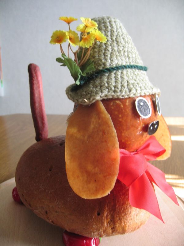 Der Brotdackel hat einen Hut mit Blumen und einen bunten Schal, sitzt dekorativ auf einem Tisch.