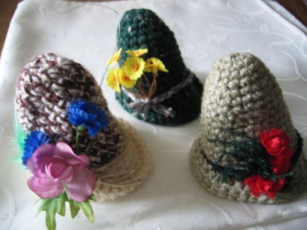 Drei gehäkelte Hüte mit bunten Blumen stehen dekorativ auf einem weißen Untergrund, ideal für ein originelles Geschenk.
