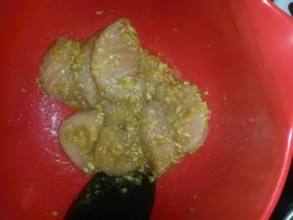 Fleischstücke mariniert in einer gelben Sauce liegen in einer roten Schüssel. Ein schwarzer Löffel steckt darin.