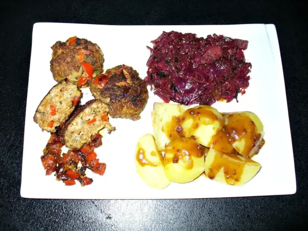 Leckere Frikadellen mit Gemüse liegen auf einem weißen Teller, umgeben von Rotkohl und mit glasierten Kartoffeln.