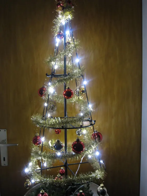 Ein künstlicher Weihnachtsbaum aus Metall leuchtet mit Lichtern und bunten Ornamenten vor einer Holzchalttür.