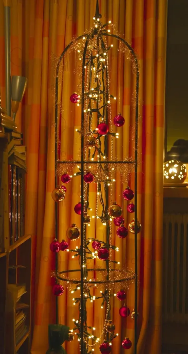 Ein dekorativer Weihnachtsbaumersatz steht im Raum, geschmückt mit Lichtern und roten Kugeln vor einem orangefarbenen Vorhang.