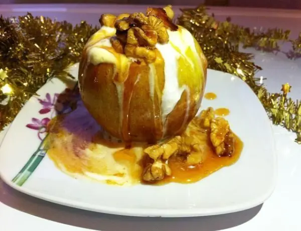 Ein Apfel, gefüllt mit Eis und karamellisierten Walnüssen, steht auf einem Teller und wird von festlichem Goldschmuck umgeben.