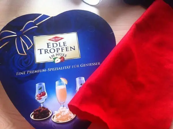 Eine herzförmige Verpackung aus Pappe mit süßen Leckereien liegt neben einem roten Tuch auf einem Tisch.