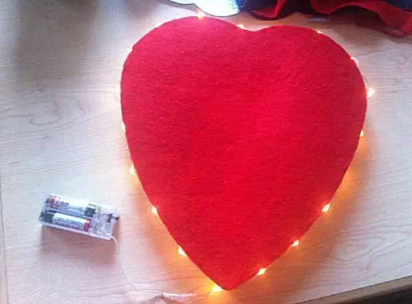 Ein rotes Herz-Kissen mit Lichtern liegt auf einem Tisch, daneben eine Batterie, um die Wand- und Dekoleuchte zu betreiben.