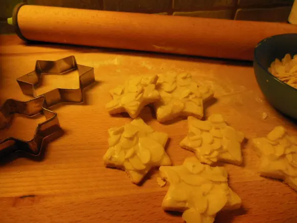 Butter-Mandel-Sterne werden ausgestochen und liegen auf einer Holzplatte, während ein Nudelholz und eine Schüssel im Hintergrund sichtbar sind.