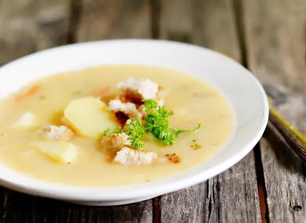 In einer weißen Schüssel liegt cremige Kartoffelsuppe, garniert mit Brotkrumen und frischer Petersilie, auf einem rustikalen Holztisch.