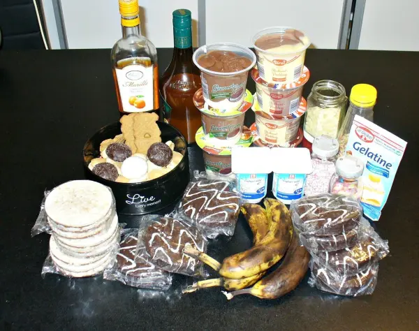 Ein Glaskuchen wird aus verschiedenen Zutaten, wie Marmelade, Bananen und Keksen, auf einem Tisch zusammengestellt.