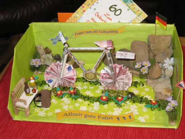 Ein dekoriertes Geldgeschenk in Form eines Fahrrads, umgeben von kleinen Steinen und Blumen, vermittelt kreatives Basteln.