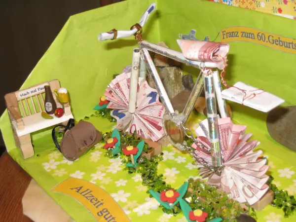 Ein aus Geldscheinen gefaltetes Fahrrad steht auf einer grünen Unterlage mit Blumen und einer kleinen Bank.