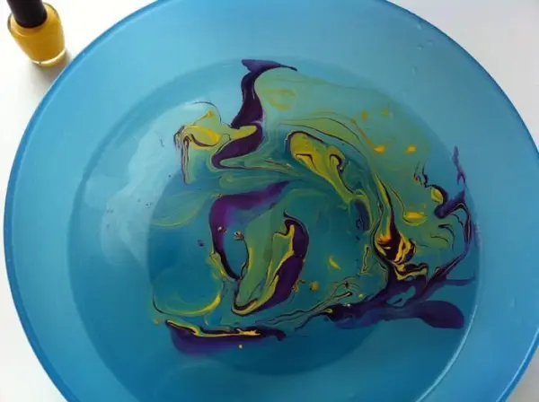 Eine blaue Schüssel mit buntem, marmoriertem Nagellack auf der Oberfläche steht neben einer Nagellackflasche.