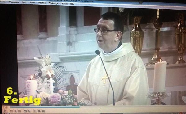 Ein Priester steht in der Kirche und spricht, während Kerzen und Blumen im Hintergrund zu sehen sind.