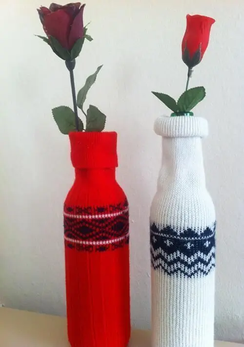 Rote und weiße Flaschen mit Blumen in gestrickten Pullovern stehen dekorativ auf einem Tisch in einem winterlichen Raum.