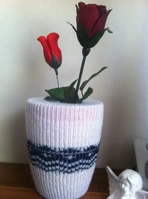 Eine dekorative Vase, in einem gestrickten Winterüberzug, hält rote Rosen und steht auf einem Tisch neben einer Figur.