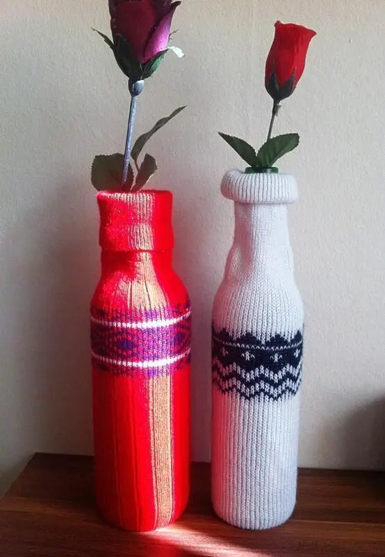 Zwei mit Stricküberzügen verzierte Flaschen stehen dekorativ auf einem Tisch, jeweils mit einer Rose oben.