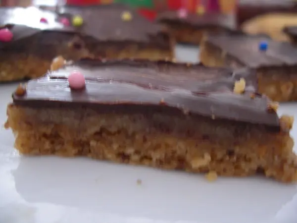 Ein Schoko-Karamell-Kuchenstück liegt auf einem Teller, garniert mit bunten Streuseln, vor einer unscharfen Hintergrunddekoration.