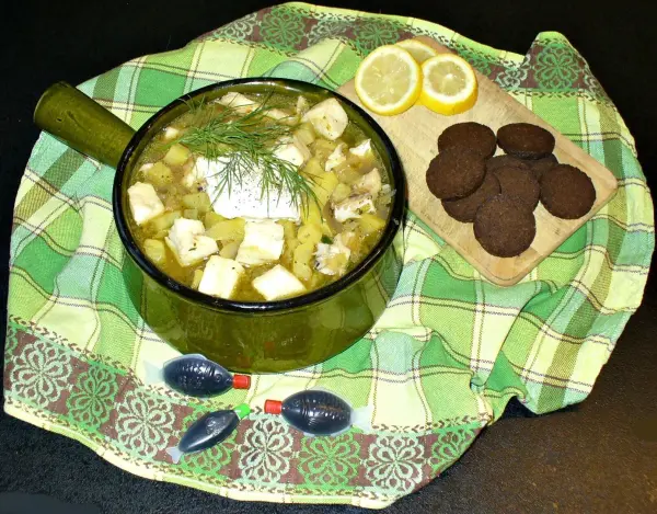 Ein grüner Topf mit schwedischem Fischtopf steht auf einem karierten Tuch, umgeben von Zitronenscheiben und Keksen.