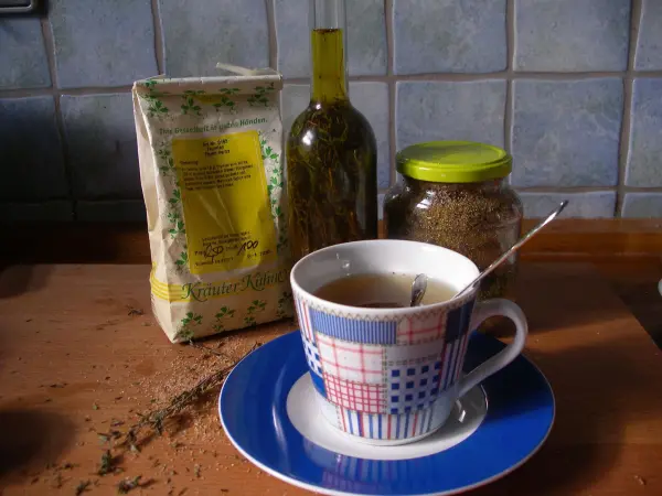 Eine Tasse Thymiantee steht auf einem blauen Unterteller, umgeben von einer Packung Thymian, einem Öl und Gewürzen auf einer Holztischplatte.