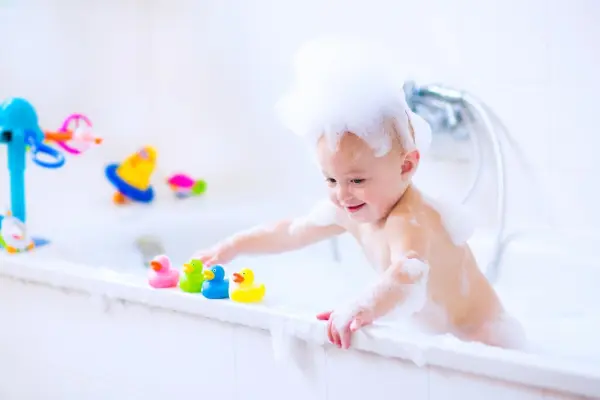Ein Kleinkind plantscht mit Spielzeugente in der Badewanne, umgeben von bunten Badewannen-Spielzeugen und Schaum.