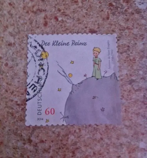 Eine Briefmarke zeigt den kleinen Prinzen, der auf einem Hügel steht, umgeben von Sternen und einer kreativen Illustration.