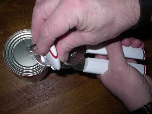 Ein Handdosenschneider öffnet eine Dose, während sich die Hand eines Benutzers auf dem Griff befindet. Ein Holzuntergrund ist sichtbar.