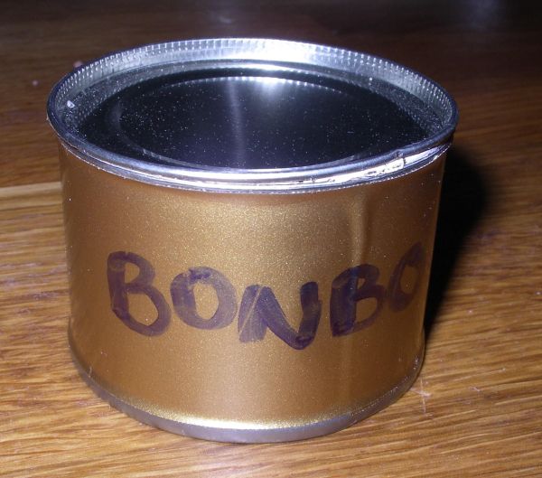 Eine goldene Dose steht auf einem Holztisch und zeigt eine aufgeschriebene Beschriftung „BONBON“. Die Dose hat keinen scharfen Rand.