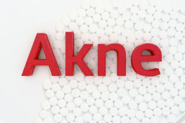 Roten Buchstaben aus "Akne" stehen auf einer weißen Hintergrundleiste mit kleinen weißen Tabletten, die den Fokus auf Hautprobleme lenken.