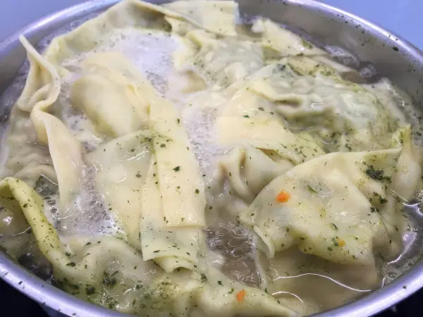 In einem Topf kochen Maultaschen in einer Brühe, umgeben von sprudelndem Wasser und Kräutern.