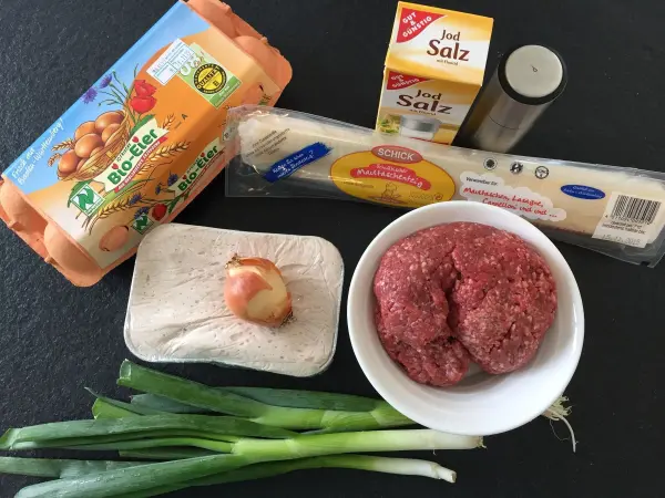 Maultaschen-Zutaten liegen auf einer schwarzen Unterlage: Eier, Hackfleisch, Zwiebeln, Brot und Salz.