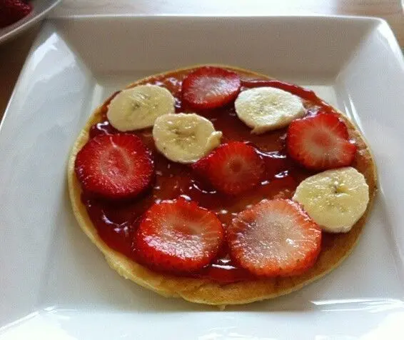 Ein Pfannkuchen liegt auf einem weißen Teller, belegt mit frischen Erdbeeren und Bananenscheiben, umgeben von einer hellen Küche.