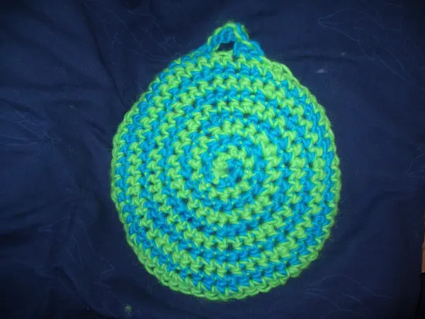 Ein gefilzter Topflappen in zweifarbigen Spiralen liegt auf einem dunklen Hintergrund und zeigt ein fröhliches Muster in Blau und Grün.