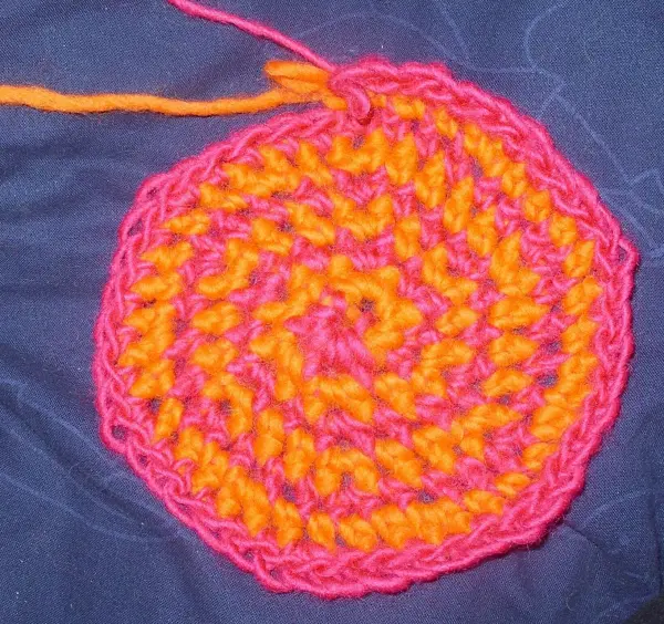 Ein zweifarbiger, gehäkelter Untersetzer in Orange und Pink liegt auf einem blauen Hintergrund, bereit zum Filzen.