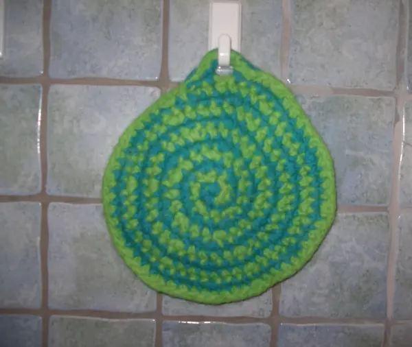 Ein gefilzter Topflappen in spiralförmigem Design hängt an einem Haken, umgeben von fliesenartigen Wänden.