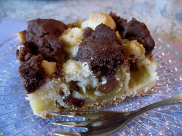 Ein Stück Apfelkuchen mit Dinkelmehl und marmorierten Streuseln liegt auf einem gläsernen Teller, umgeben von einem hellen Hintergrund.